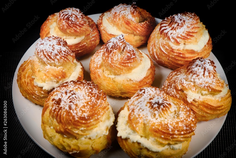 Buns choux pastry. Cakes sprinkled with powdered sugar. Scones with custard on a plate. Bun with sweet cottage cheese. Dark background.