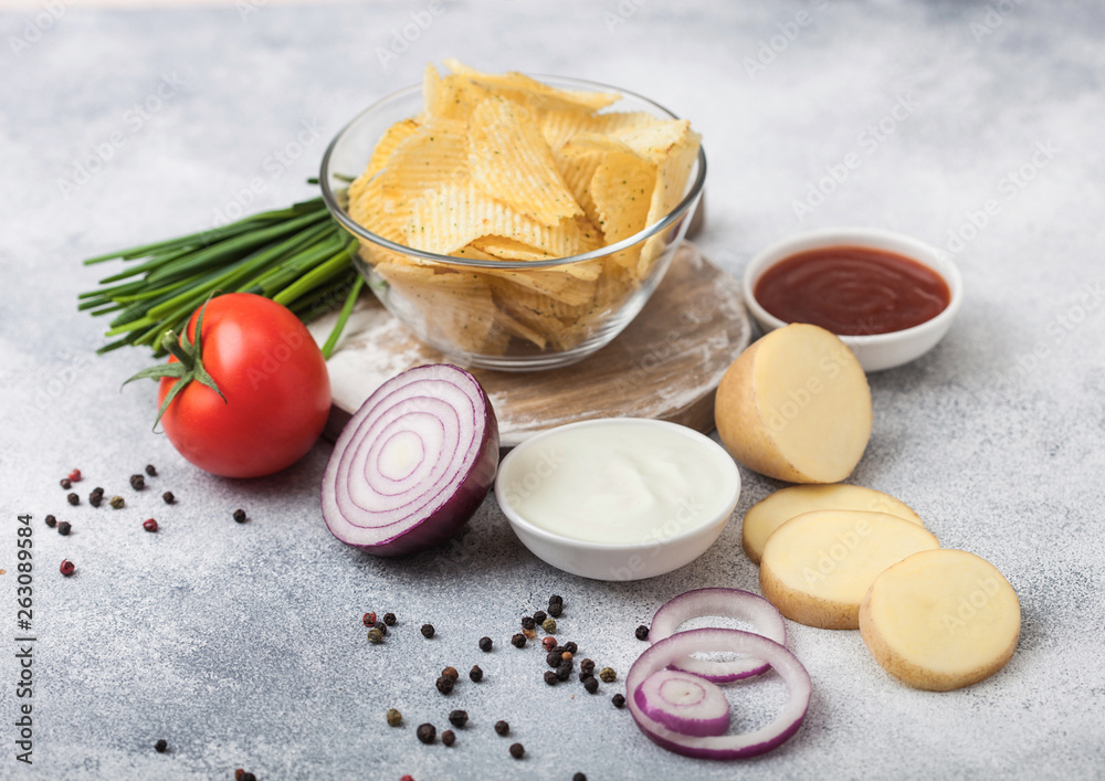 Fresh organic homemade potato crisps chips in glass bowl with sour cream and red onions on light kitchen table background. Tomato with ketchup and green onion