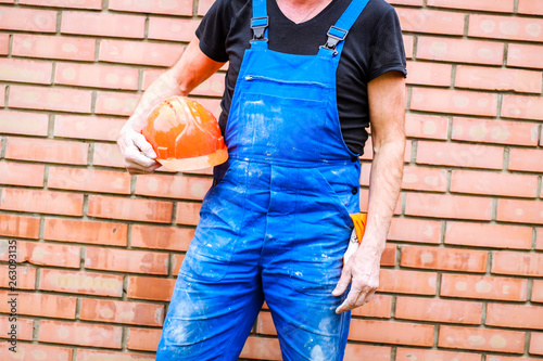 A builder with a construction cup in his hands against the background of a brick wall. Blue form combining.