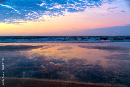 The beautiful soft pinks and blues of early sunrise at the beach are reflected on ripples in the water at low tide.