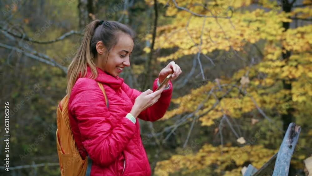 pretty woman hiker in red jacket with small backpack is standing on the yellow autumn woods background using phone and taking photo of autumn landscape. pretty woman hiker in red jacket with small backpack is standing on the yellow autumn woods background using phone and taking photo of autumn landscape.