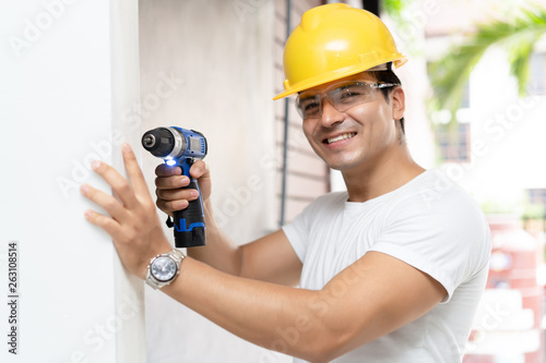 Man in helmet with electric drill making hole in wall and looking at camera. Architecture and Home renovation concept.