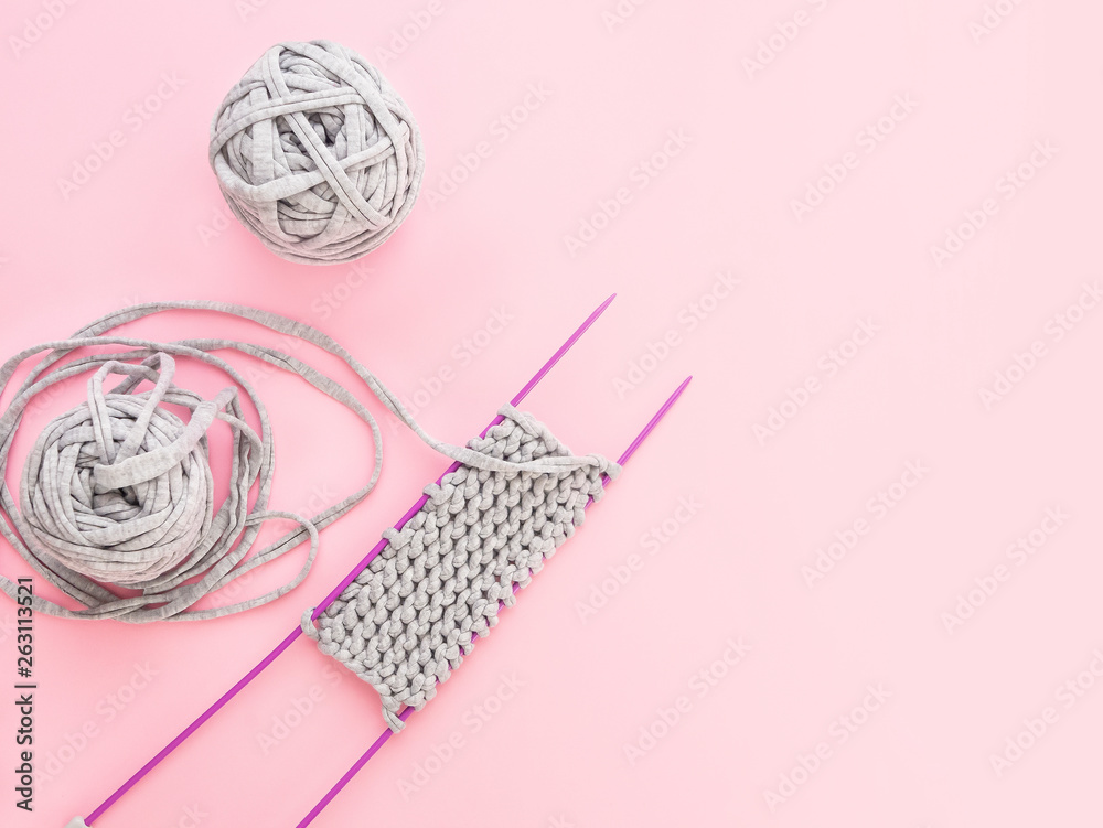 Knitting project in progress. A piece of knitting with ball of grey yarn and a violet knitting needles on pink bacground. Hand made concept, hobby for rest, textile idea.Top view.Copy space Knitting project in progress. A piece of knitting with ball of grey yarn and a violet knitting needles on pink bacground. Hand made concept, hobby for rest, textile idea.Top view.Copy space