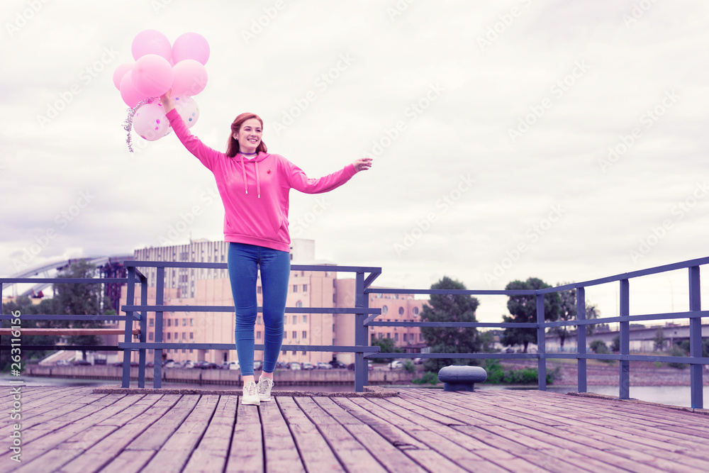 Beaming ginger woman walking on wooden pier with balloons
