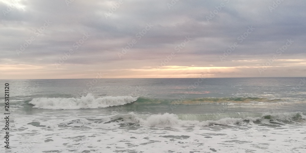 Sunset seascape. Autumn storm sea. Gray, blue, turquoise waves with white foam break on a rocky beach. The sun breaks through the low heavy clouds. Background