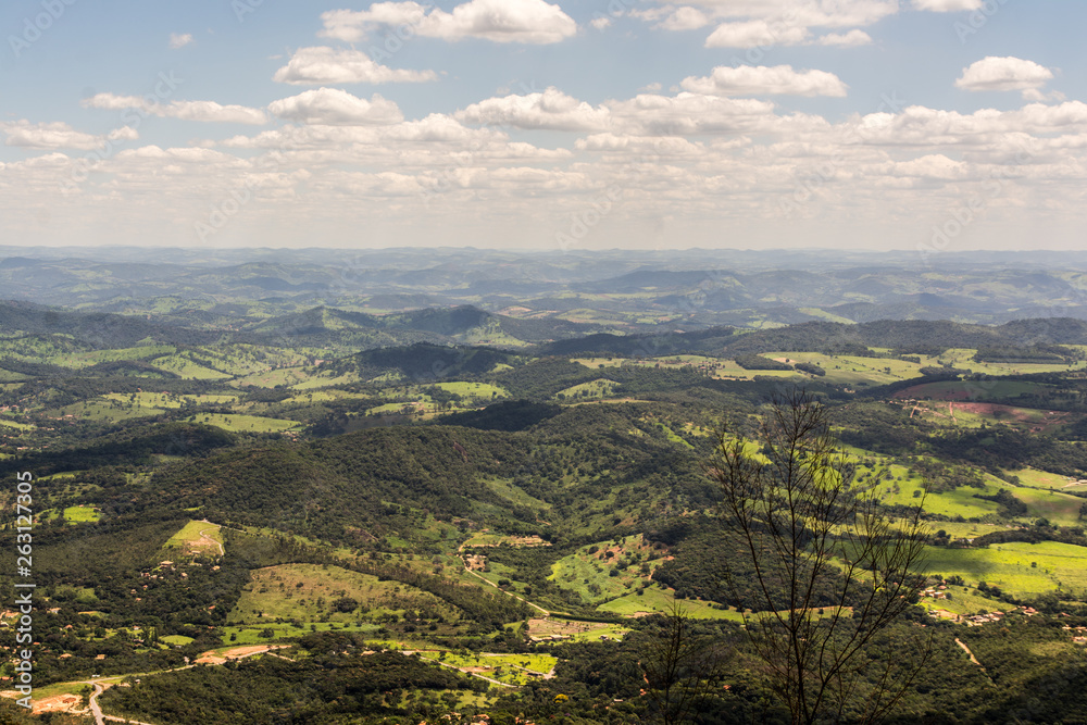Aerial view of green hilly landscape in rural Brazil. A twig is seen on the background, and sparsely clouded skies project shadows along the hills.