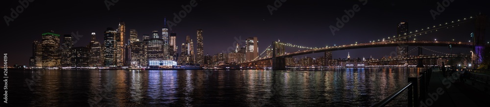 A large panoramic nighttime view of Manhattan and the Brooklyn Bridge from the opposite river bank