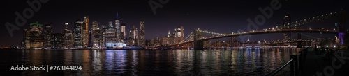 A large panoramic nighttime view of Manhattan and the Brooklyn Bridge from the opposite river bank