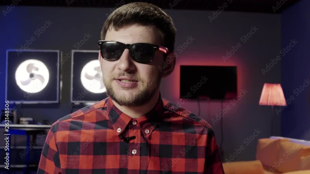 Young cute white bearded man vlogger put on stylish sunglasses and red checkered shirt with small wireless mic speaks at camera and actively claps in room modern furniture. Young cute white bearded man vlogger put on stylish sunglasses and red checkered shirt with small wireless mic speaks at camera and actively claps in room modern furniture.