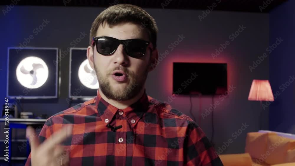 Cool cute caucasian bearded guy wearing stylish sunglasses and red checkered shirt with small wireless mic looks at camera and emotionally speaks in modern furnished room. Cool cute caucasian bearded guy wearing stylish sunglasses and red checkered shirt with small wireless mic looks at camera and emotionally speaks in modern furnished room.