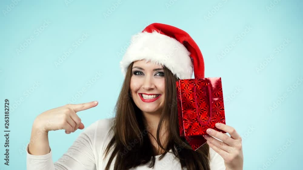 Happy young woman in santa claus hat holds small red shopping bag. Girl received gift, opens it and rejoices, on blue. People celebrating xmas love and happiness concept Happy young woman in santa claus hat holds small red shopping bag. Girl received gift, opens it and rejoices, on blue. People celebrating xmas love and happiness concept
