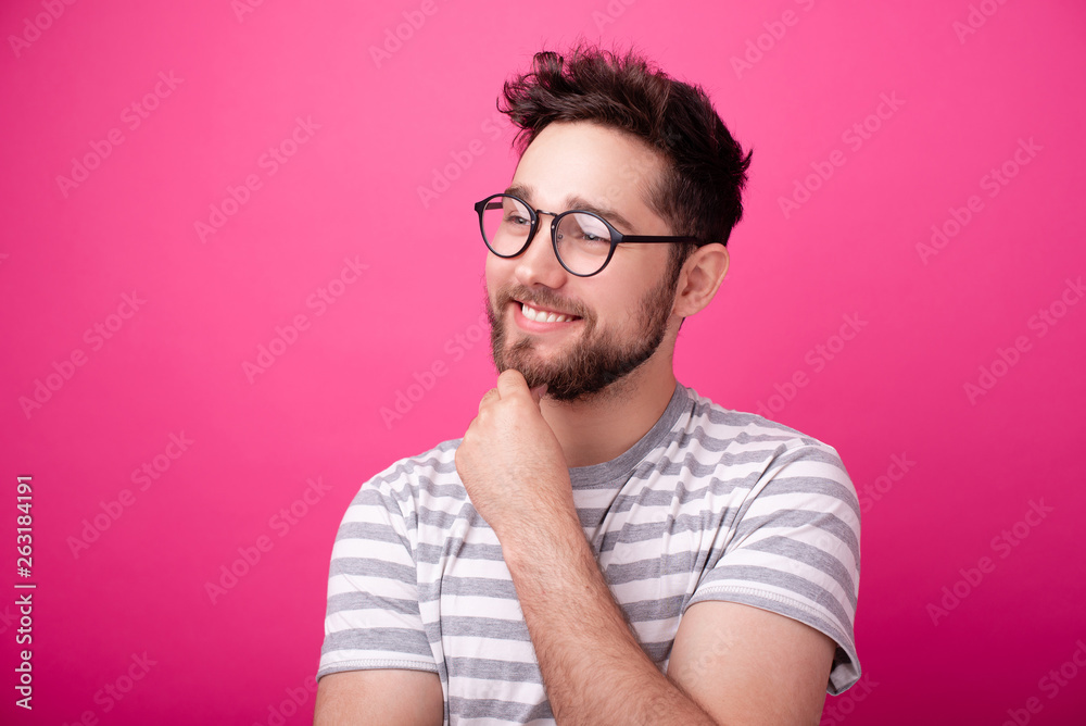 Fototapeta premium Handsome young bearded man wearing glasses, looking far away and touching his chin over pink background