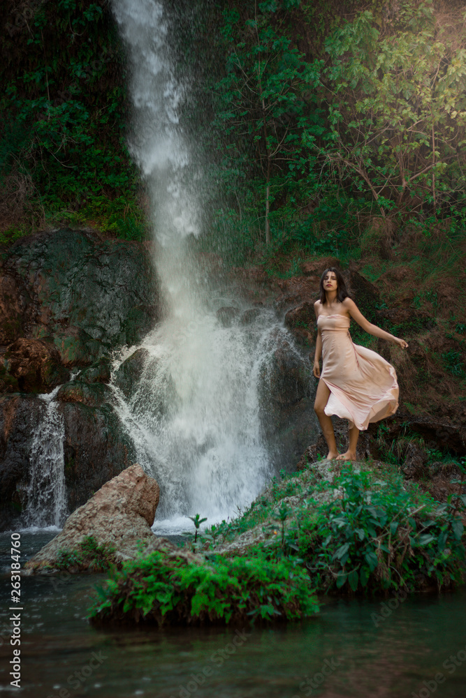 Obraz premium Sexy woman posing on the rock among green tropical plants and beside beautiful waterfall with blue water. Fairy tale.
