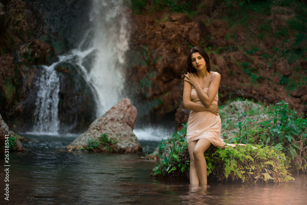 Sexy woman posing on the rock among green tropical plants and beside beautiful waterfall with blue water. Fairy tale.