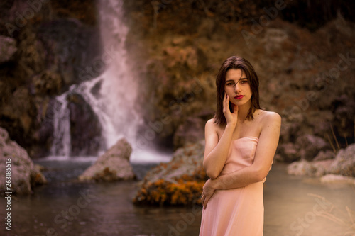 Attractive girl posing on the rock among in fairy fast forest in autumn and beside beautiful waterfall with blue water. Fairy tale.