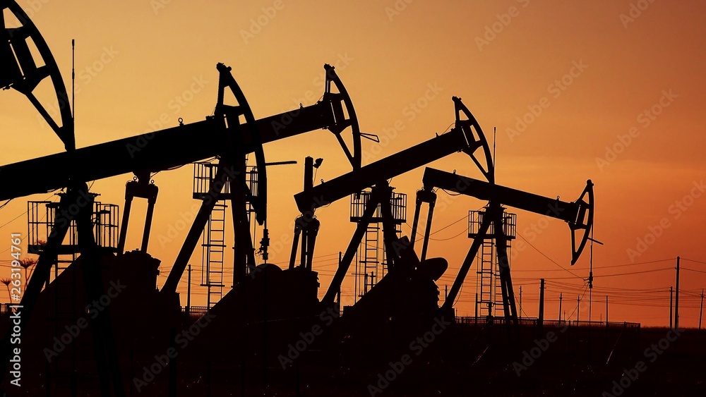 Many oil pumps at sunset under the red sky on industrial platform field with crude petroleum hydraulic extraction units