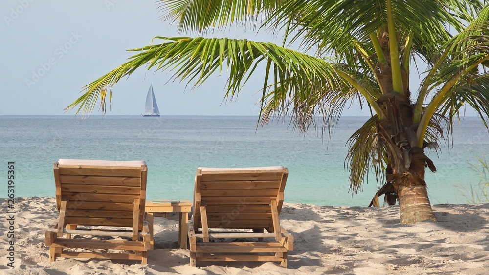 Wooden lounge sun beds under coconut palm tree on exotic beach and boat sailing away on sea Wooden lounge sun beds under coconut palm tree on exotic beach and boat sailing away on sea
