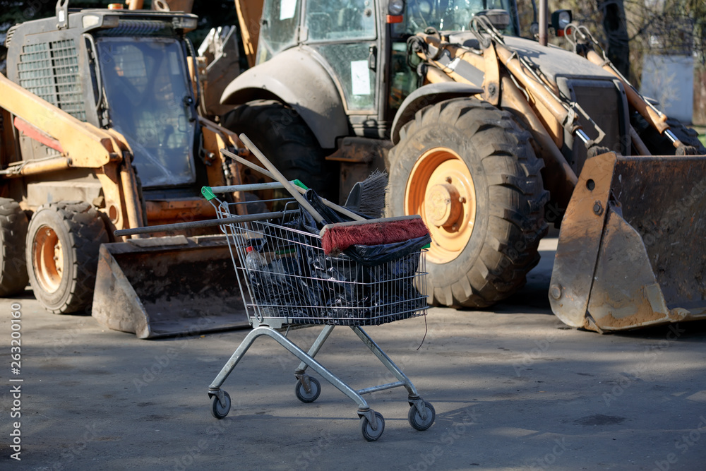 trolley from the supermarket filled with cleaning items, brooms and mops on the background of harvesting equipment