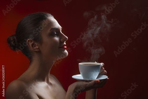 Attractive lady brunette with pleasure holding a coffee cup with light smoke in hand in studio at the red background
