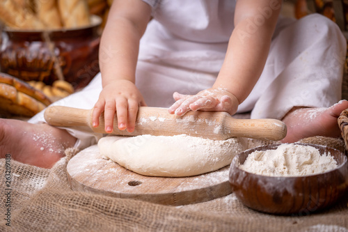 a small child sits on a wooden table and rolls out the dough with a rolling pin,flour is scattered around and bread lies. the child develops fine motoriku gets skills