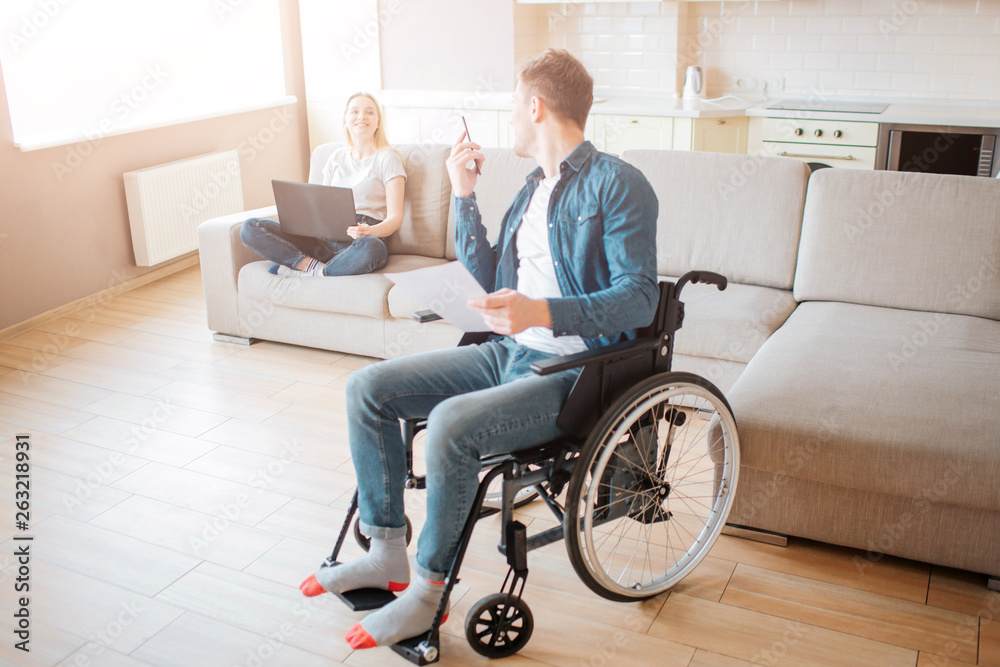 Cheerful couple in room together. Young man with inclusiveness sit on wheelchair and look back on woman. Holding phone in hands nad laptop on knees. Smile to each other. Cheerful couple in room together. Young man with inclusiveness sit on wheelchair and look back on woman. Holding phone in hands nad laptop on knees. Smile to each other.