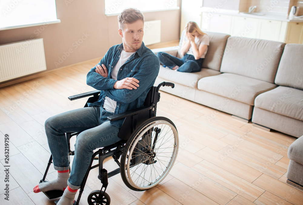 Young man with inclusiveness and disability sit on wheelchair in front. Hands crossed and upset. Young woman sit behind on sofa and cry. Emotional sickness and stress. Young man with inclusiveness and disability sit on wheelchair in front. Hands crossed and upset. Young woman sit behind on sofa and cry. Emotional sickness and stress.