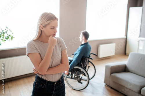 Young man sit with special needs sit on wheelchair and look on window. Woman stand in front and think. Argue and quirrel. Couple upset and unhappy.