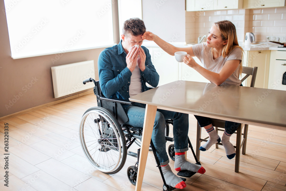 Sick young man with special needs and disability sneezing. Sick and ill guy. Young woman sit beside in kitchen and take care of him. Daylight.