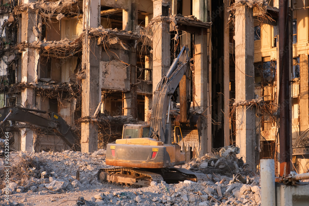 Fototapeta premium Building demolition with hydraulic excavator. Dismantle of destructed house ruins at bright sunsest