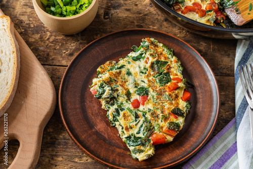 A piece of frittata with spinach and red pepper on a plate and a cutting board with bread, rustic style, top view photo