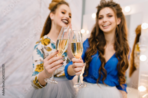 Two glasses of champagne holding by attractive happy young women having fun in hairdresser salon. Expressing true positive emotions  smiling  chilling  luxury lifestyle  fashionable models