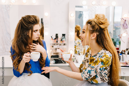 Two astonished attractive women talking in beauty salon. Drinking coffee, gossip, preparing to party, having fun, friends together, true emotions, professional makeup, coiffure photo