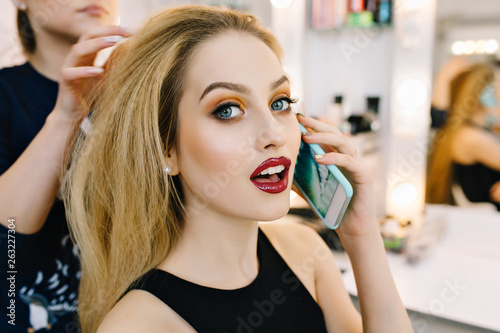Closeup portrait gorgeous blonde model preparing to celebration, party in hairdresser salon. Stylish makeup, making hairstyle, talking on phone, looking to camera, luxury look