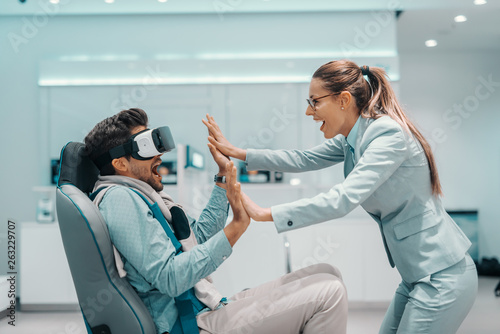 Amazed mixed race man sitting in chair and trying out vr technology. Woman standing in front of man and holding his hands. Tech store interior.