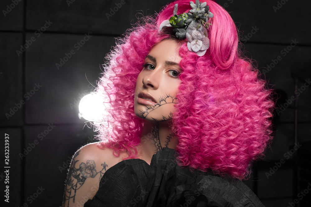 Fototapeta premium young attractive caucasian girl model with afro style curly bright pink hair, tattooed face and flowers woven into her hair. Photo in the studio on a black background