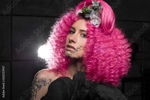 young attractive caucasian girl model with afro style curly bright pink hair, tattooed face and flowers woven into her hair. Photo in the studio on a black background