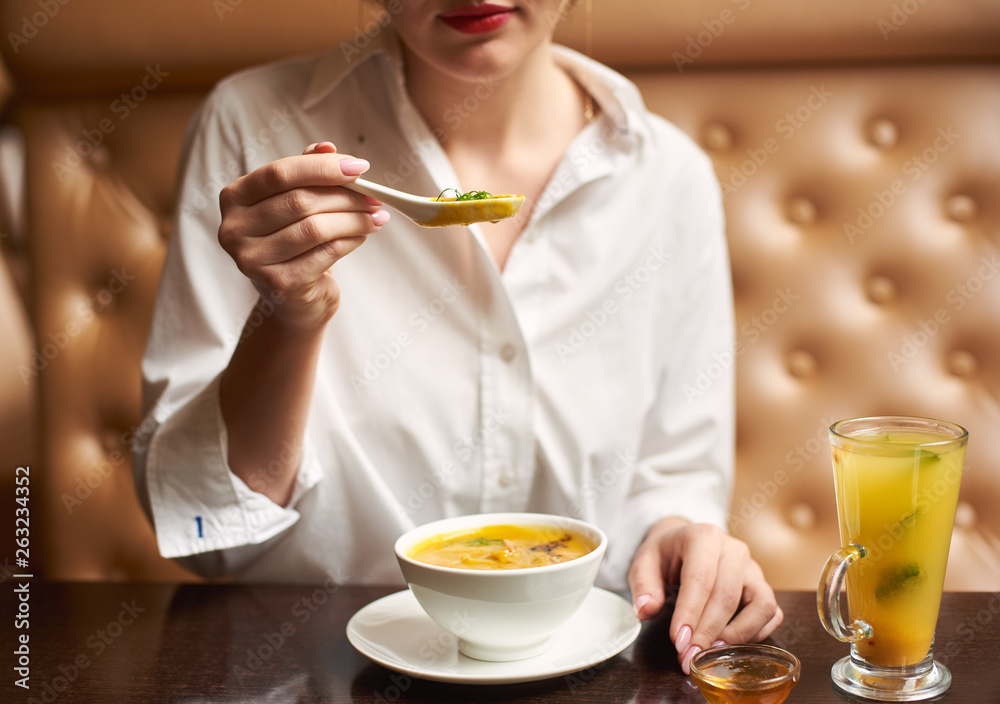 Crop of beautiful girl eating tasty orange cream soup. Woman in white blouse enjoying tasty first dish in luxury restaurant. Client eating exotic meal on lunch. Concept of oriental food. Crop of beautiful girl eating tasty orange cream soup. Woman in white blouse enjoying tasty first dish in luxury restaurant. Client eating exotic meal on lunch. Concept of oriental food.