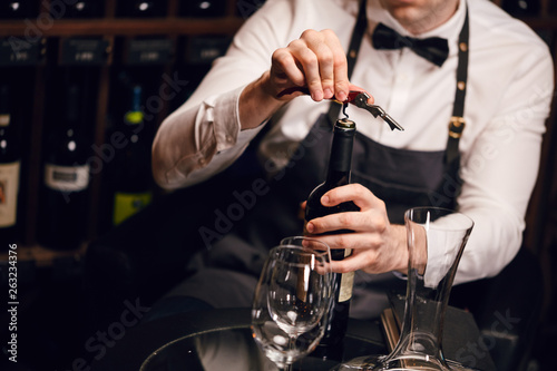 Elegant young sommelier with bow tie uncorking bottle of wine in wine boutique. Wine tasting social event. photo