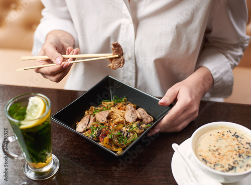 Woman ordering oriental style meal in restaurant. Client enjoying lunch including soup with seeds, warm salad with meat and lemon mint tea. Close up hands with manicure holding food sticks.
