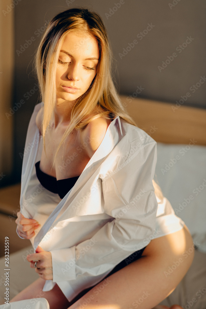 Young blonde woman sitting on unmade bed, looking really sexy with shirt  down on her bare shoulder. Stock Photo | Adobe Stock