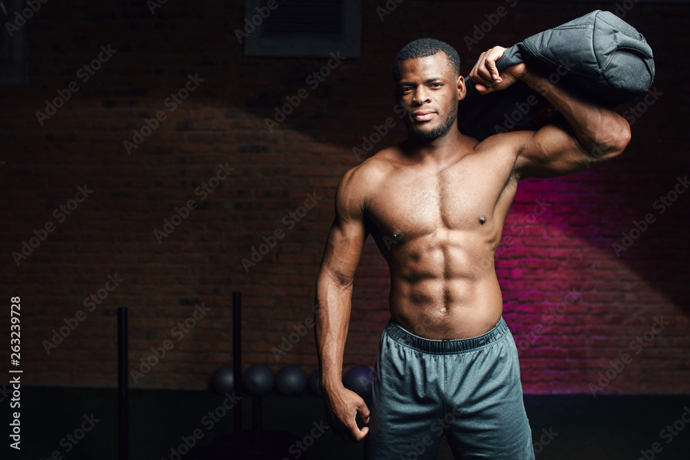 Cross fit male athlete with naked muscular torso doing exercise with cross fit sports equipment, raising heavy sandbag as a dumbbell over dark background