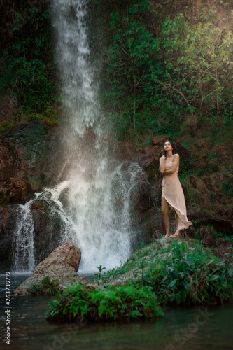 Sexy woman posing on the rock among green tropical plants and beside beautiful waterfall with blue water. Fairy tale.