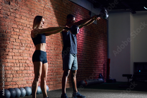 Young caucasian woman athlete working out with kettlebells with the help of personal trainer during body core cross fit workout