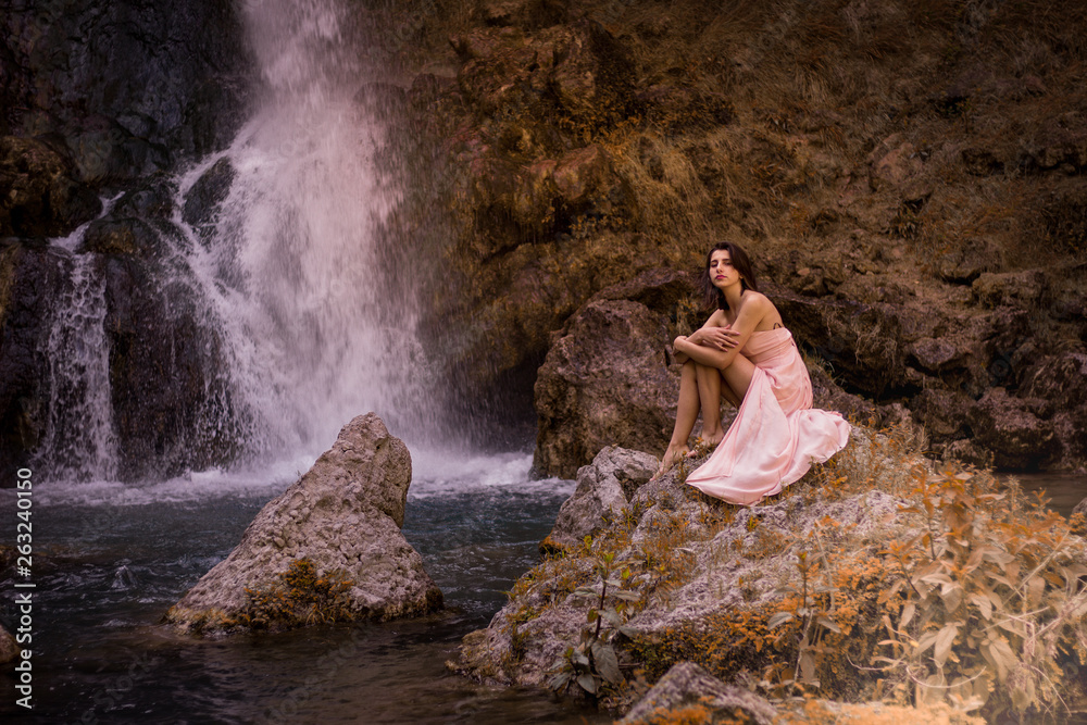 Fototapeta premium Attractive girl posing on the rock among in fairy fast forest in autumn and beside beautiful waterfall with blue water. Fairy tale.
