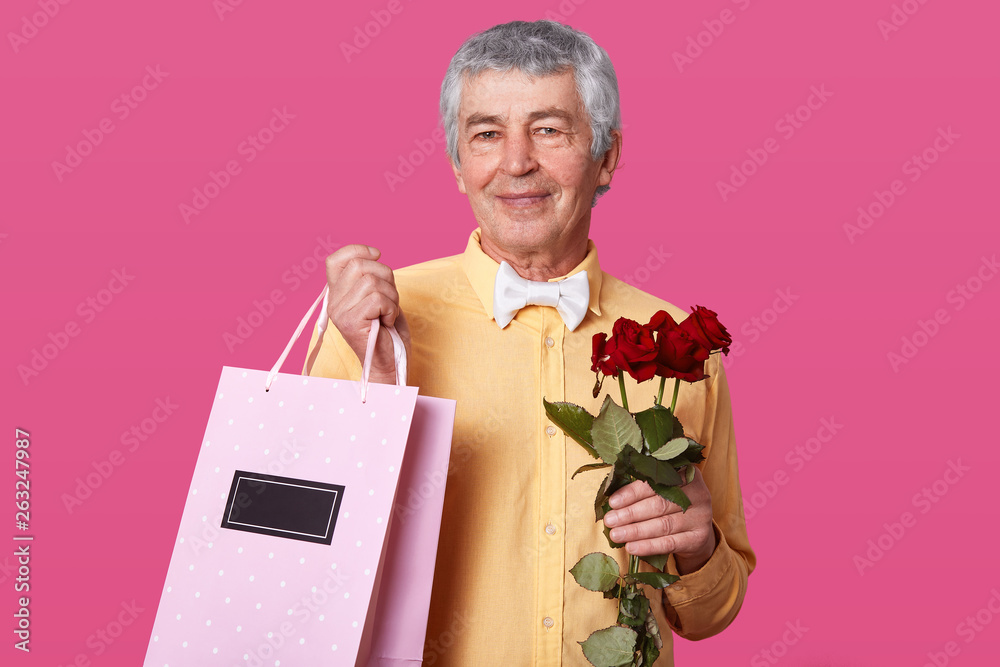 Photo of attractive mature man with pleasant facial expression, dressed in yellow shirt with white bowtie, carries pink bag with present and roses, wants to congratulate wife with wedding anniversary. Photo of attractive mature man with pleasant facial expression, dressed in yellow shirt with white bowtie, carries pink bag with present and roses, wants to congratulate wife with wedding anniversary.