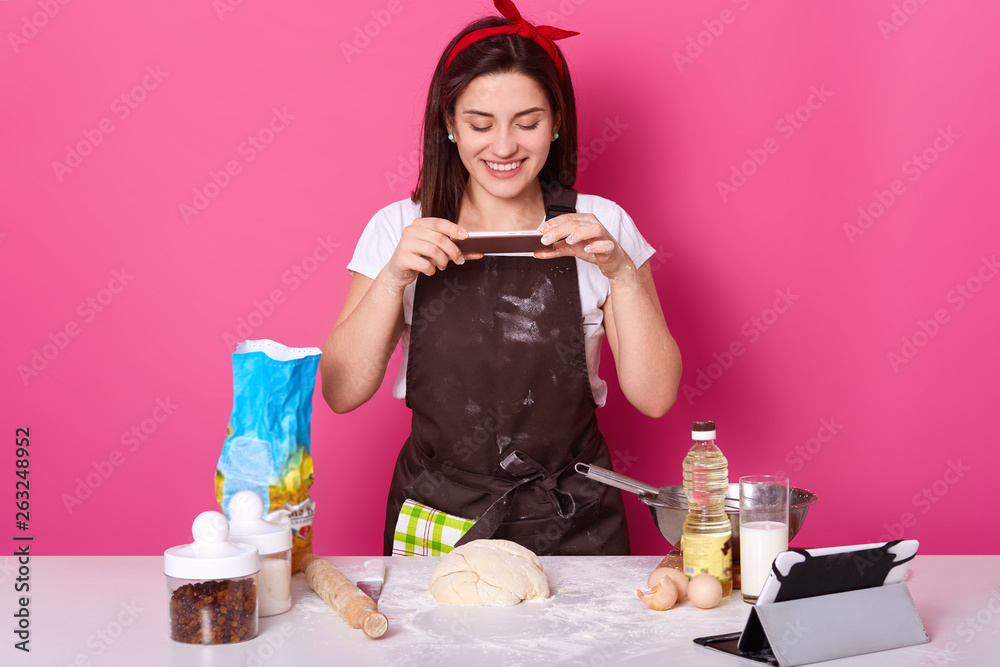 Young European pleasant woman makes photo of her half ready pie, post it in social networking sites. Cheerful attractive cute model poses in brown apron, white t shirt, and bright red headband. Young European pleasant woman makes photo of her half ready pie, post it in social networking sites. Cheerful attractive cute model poses in brown apron, white t shirt, and bright red headband.