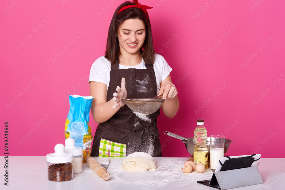 Portrait of skillful talented cook putting flour through sieve on half ready pie with raisin. Brunette cute young model poses isolated over bright pink background. Baking and cooking concept. Portrait of skillful talented cook putting flour through sieve on half ready pie with raisin. Brunette cute young model poses isolated over bright pink background. Baking and cooking concept.