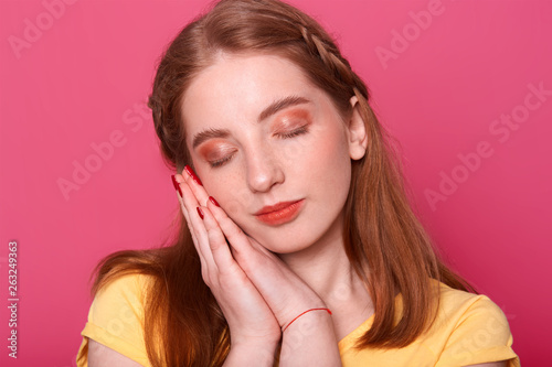 Indoor studio shot of romantic pretty young lady with closed eyes and hands near face, going to have nap, looks sweet. Attractive model poses isolated over bright pink background. Youth concept.