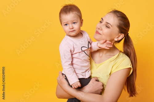 Studio shot of loving mother holding her baby girl, toddler looking aside, mom admires her charming doughter isolated over yellow studio wall. Motherhood, happyness, children concept. Copy space.