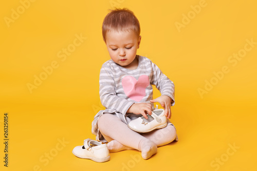 Fair haired pretty little child wearing stripped dress with heart poses isolated over bright yellow background. Sweet magnetic kid sits on floor in studio holding and observing her white shoes.
